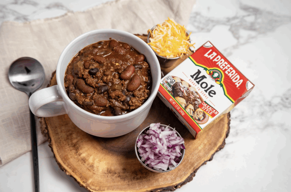 A hearty bowl of Mole Turkey Taco Chili made with La Preferida Mole Sauce, kidney and black beans, topped with shredded cheese and diced red onions on the side.