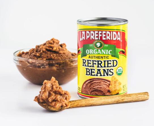 Can of La Preferida Organic Authentic Refried Beans next to a bowl and spoon of prepared refried beans ready to heat and serve.