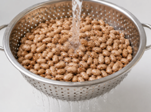 Dry pinto beans being rinsed under running water in a colander before soaking and cooking.